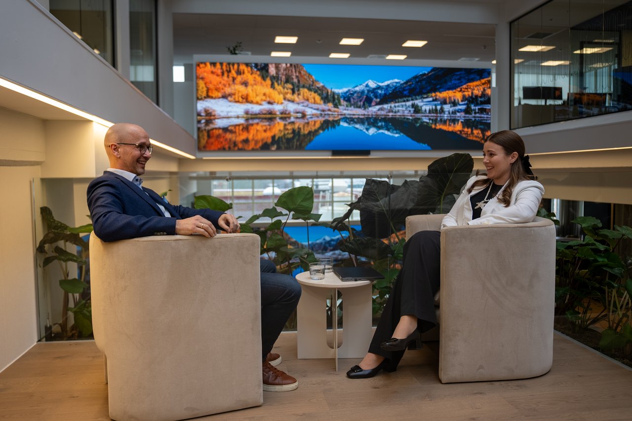Two colleagues meeting in a modern office lounge space featuring digital signage and collaborative work areas.
